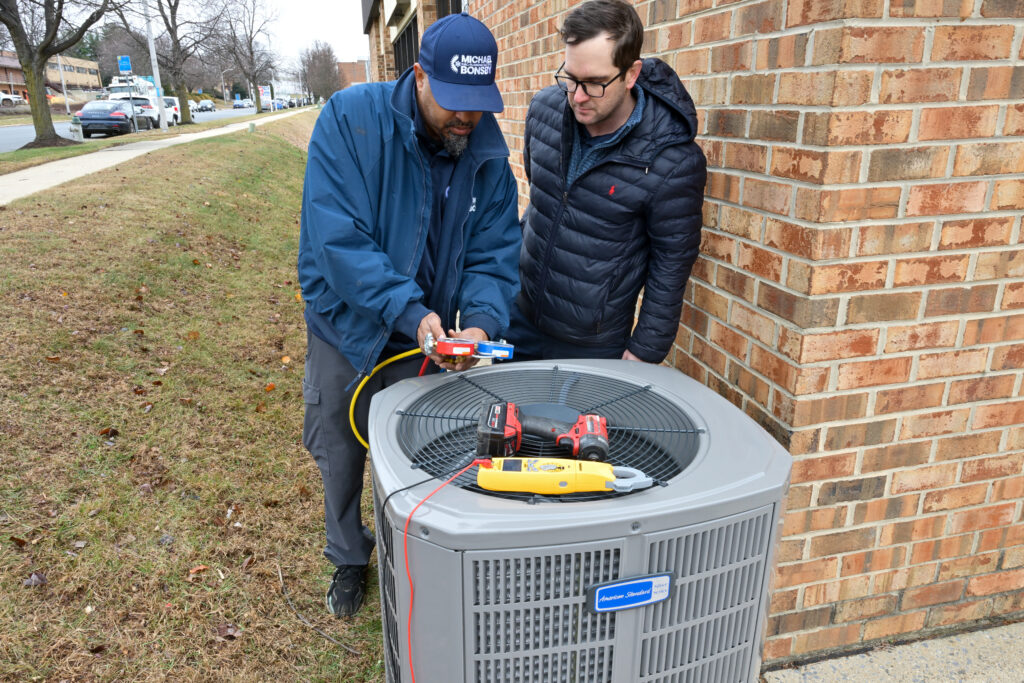 An HVAC technician in a blue "Michael Bonsby" uniform and hat explains a set of pressure gauges to a customer. They are standing outdoors next to a grey American Standard air conditioning unit. Several tools, including a red power drill and a yellow multimeter, are resting on top of the unit's metal grate.