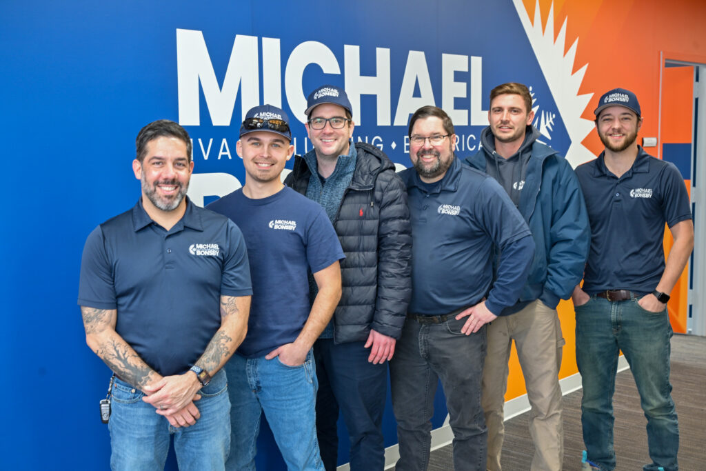 A group of six men, likely employees of Michael Bonsby HVAC, Plumbing & Electrical, posing for a photo in front of a blue and orange company logo wall. They are wearing navy blue company-branded apparel, including polo shirts, t-shirts, jackets, and baseball caps. The team is smiling and standing in a casual line against the vibrant backdrop.
